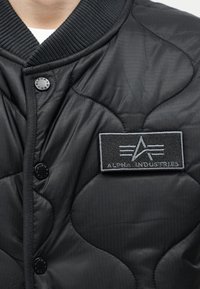 Μαύρο καπιτονέ μπουφάν με ριμπ γιακά, κουμπιά με πατιλέτα και γκρι κεντημένο έμβλημα "Alpha Industries" στο στήθος.