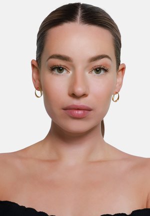 Goldene Creolen mit strukturiertem Finish, kreisförmigem Design und glatten Kanten, getragen von einer Person mit neutralem Make-up vor einem schlichten Hintergrund.