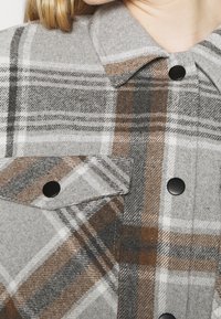 Gray plaid shirt made of wool blend, featuring brown, white, and black stripes, with a buttoned collar and flap pockets.