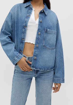 Mujer con chaqueta de mezclilla azul claro sobre top blanco corto y jeans de mezclilla azul claro a juego, con la mano apoyada en la cintura del jeans.