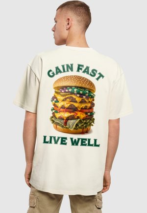 Krémovo zafarbené oversized tričko s veľkým grafickým motívom hamburgera na chrbte, s zeleným textom "GAIN FAST" a "LIVE WELL."