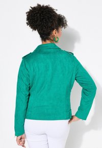 Giacca in camoscio verde con colletto, spalline, maniche lunghe e una forma aderente. Superficie testurizzata e dettagli minimi.