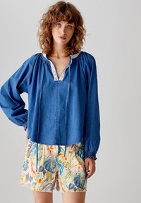Top in denim blu con maniche lunghe, spalle a sbuffo e rifinitura bianca. Abbinato a pantaloni corti floreali luminosi e a fantasia con vari disegni di foglie.