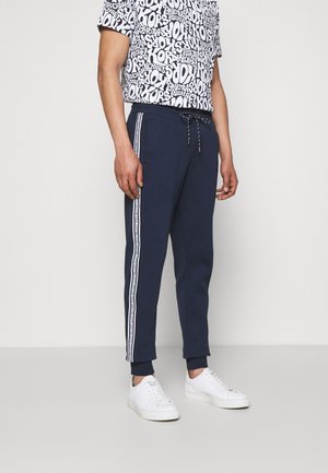 Man draagt marineblauwe joggers met witte strepen aan de zijkant en een zwart-wit grafisch T-shirt, gecombineerd met witte sneakers, staand tegen een witte muur.