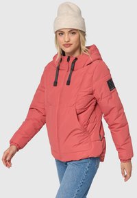 Polstret koraljakke med lynlås, sorte snøre, store lommer og logo-patch på ærmet. Bæret med blå jeans og en lys beige hue.