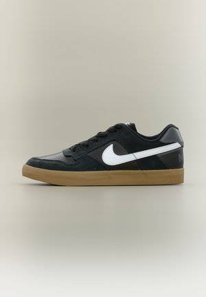 Baskets basses noires avec le logo Nike swoosh blanc, lacets noirs et semelle en caoutchouc marron sur un fond neutre.