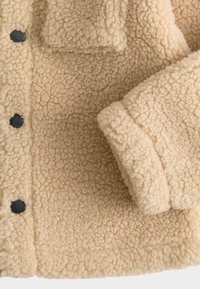 Giacca in pile beige con superficie testurizzata, dotata di bottoni neri e una tasca sul petto. Tessuto morbido con un aspetto accogliente e caldo.