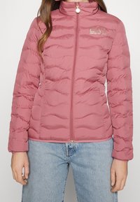 Světle růžová prošívaná bunda s vysokým límcem, zapínáním na zip, vlnitým prošíváním a malým logem na hrudi. Měkká textura tkaniny.