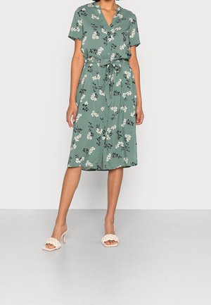 Žena v zelených midi šatech na knoflíky s krátkými rukávy, bílé květinové potisky a páskem v pase, doplněné bílými pletenými sandály na podpatku.