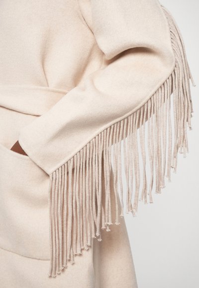 Beige fleecetröja med långa, dekorativa fransar längs ärmarna, med en omslagdesign och sidofickor för extra funktionalitet.