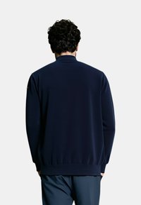 Maglione blu navy con collo alto, maniche lunghe, polsini e orlo a costine, texture liscia e dettaglio discreto del logo sulla manica.