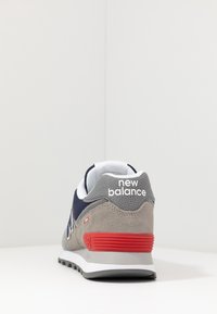 Sneaker New Balance grigio e blu navy con dettaglio rosso sul tallone, mostrato da dietro su una superficie bianca contro uno sfondo bianco uniforme.