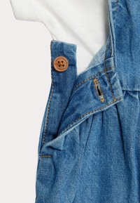 Jeans-Latzhose mit einem weißen Baumwoll-T-Shirt darunter, ausgestattet mit einem braunen Knopf, blauen Nähten und einem gefältelten Design in der Taillengegend.