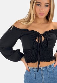 Schwarzes Off-Shoulder-Crop-Top mit rüschengeschnittenen Kanten, Bindeband-Detail und gerafftem Textil; lange, voluminöse Ärmel. Kombiniert mit hoch sitzenden Jeans.