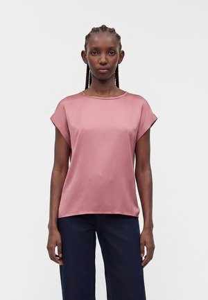 Kvinne med flette i håret som har på seg en kortermet støvete rosa bluse og mørkeblå jeans, stående foran en ensfarget lys bakgrunn.