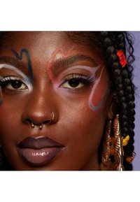 Porträtt av en person med mörk hud och färgglad ansiktsmakeup i virvlar, långa ögonfransar, djup lila läppar och stora guldörhängen med strukturerade mönster.