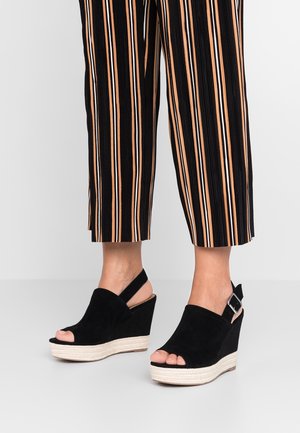 Czarne sandały na koturnie zamszowe, z odkrytymi palcami, z paskiem z tyłu i plecioną platformą z juty dla dodatkowej wysokości. Widoczne są paski spodni z podwiniętymi nogawkami.