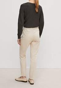Haut noir à manches longues, pantalon beige clair ajusté, texture lisse, chaussures plates avec une boucle discrète, fond uni.