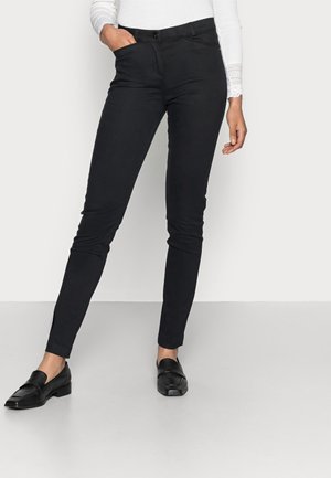 Vrouw draagt strakke zwarte broek en zwarte loafers, staat met één hand in de zak tegen een effen achtergrond.