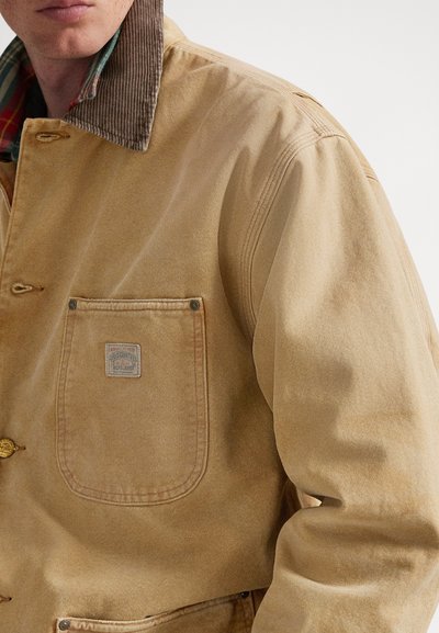 Καφέ μπουφάν εργασίας από ανθεκτικό ύφασμα, με γιακά από καφέ μουαίν, τσέπη στο στήθος με ετικέτα και κλείσιμο με κουμπιά.
