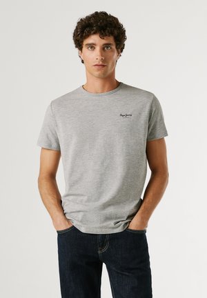 Fiatal férfi göndör hajjal, világosszürke Pepe Jeans pólót és sötét farmert viselve, zsebre tett kézzel áll egyszerű háttér előtt.