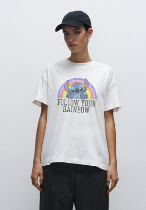 Young person wearing a black cap and white T-shirt with a colorful cartoon character and text "FOLLOW YOUR RAINBOW" on plain background.