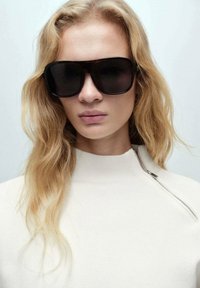Čierne oversized slnečné okuliare s elegantným rámom, nosené s krémovým topom s vysokým golierom a detailom na zips. Svetlé vlnité vlasy sú čiastočne viditeľné.