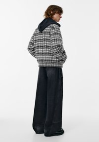 Giacca a quadri grigia e nera con cappuccio scuro, abbinata a pantaloni denim scuro a gamba larga e stivali neri, che mette in risalto una vestibilità rilassata.