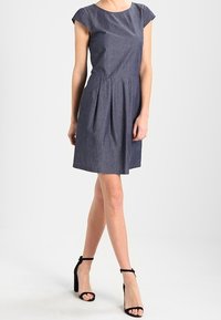 Denimkleid mit kurzen Kappenärmeln, weit ausgestelltem Rock und Faltenwurf. Dunkelblau, kombiniert mit schwarzen Riemchen-Absätzen. Glatte Textur.
