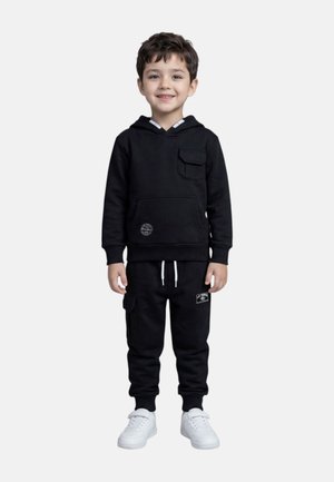 Jeune garçon souriant, portant un sweat à capuche noir avec poche avant, un pantalon de jogging noir à poche cargo, et des baskets blanches, debout.