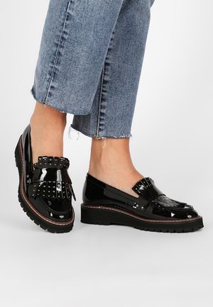 Chaussures en cuir verni noir avec une semelle plate, des franges et des détails cloutés, présentant un bout arrondi et des coutures contrastantes marron.