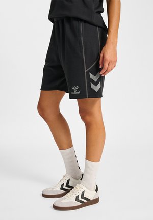 Person trägt schwarze Sportshorts mit reflektierendem Logo, weiße Socken und weiße Sneaker mit schwarzen Chevron-Mustern, steht auf einem einfarbigen Hintergrund.