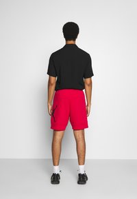 Schwarzes T-Shirt mit kurzen Ärmeln, kombiniert mit roten Sweathorts, die ein schwarzes Silhouettenlogo zeigen, in einem lässigen Schnitt und aus weichem Material.