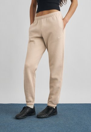 Persona con pantalones deportivos beige de Nike con puños elásticos, zapatillas negras y una camiseta negra recortada, de pie sobre un suelo alfombrado azul.