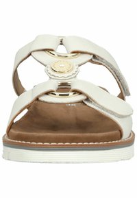 White sandal with crisscross straps, circular gold accent, and brown cushioned insole. Features a textured white sole with thin brown stripe.