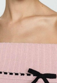 Růžový pletený top s žebrovanou texturou, zdobený černou mašlí a dekorativním řaděním černého stehu podél výstřihu.