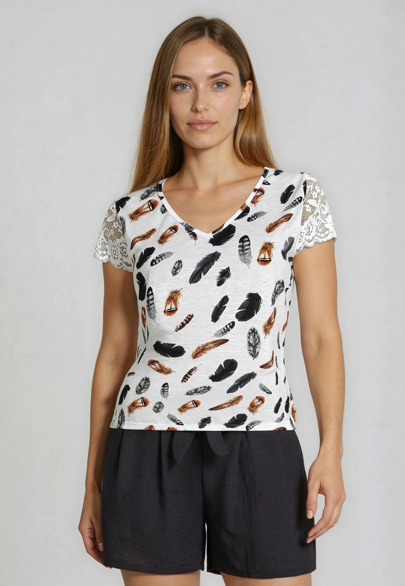 Femme aux longs cheveux portant un t-shirt blanc à col en V avec des motifs de plumes et des manches en dentelle, associé à un short noir ample.