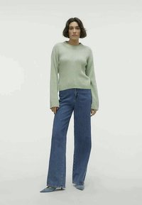 Nainen seisoo päällään vaalea vihreä ribbihuppari, leveät siniset farkut ja yksityiskohtaisilla sinisillä korkokengillä, taustanaan yksinkertainen valkoinen tausta.