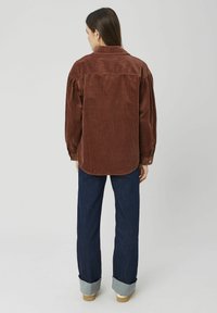 Chemise en velours côtelé marron avec un empiècement dans le dos, coupe décontractée et fermeture par boutons, associée à un jean en denim foncé aux revers retroussés.