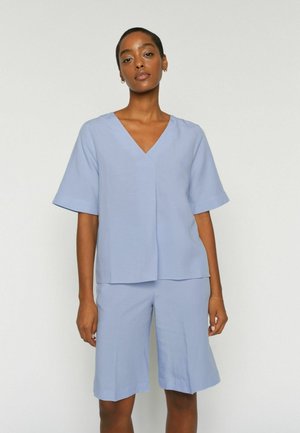 Kobieta ubrana w jasnoniebieski top z krótkim rękawem i dekoltem w serek oraz pasujące szorty do kolan, stojąca na tle jednolitej białej ściany.