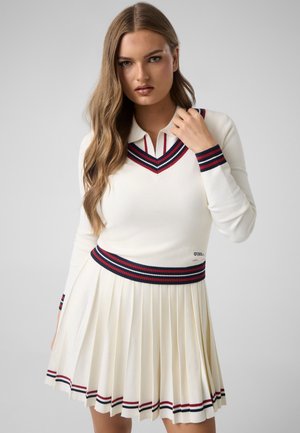 Kvinde med langt hår iført en hvid plisseret nederdel og matchende sweater med røde og marineblå striber, poserer mod en ensfarvet baggrund.