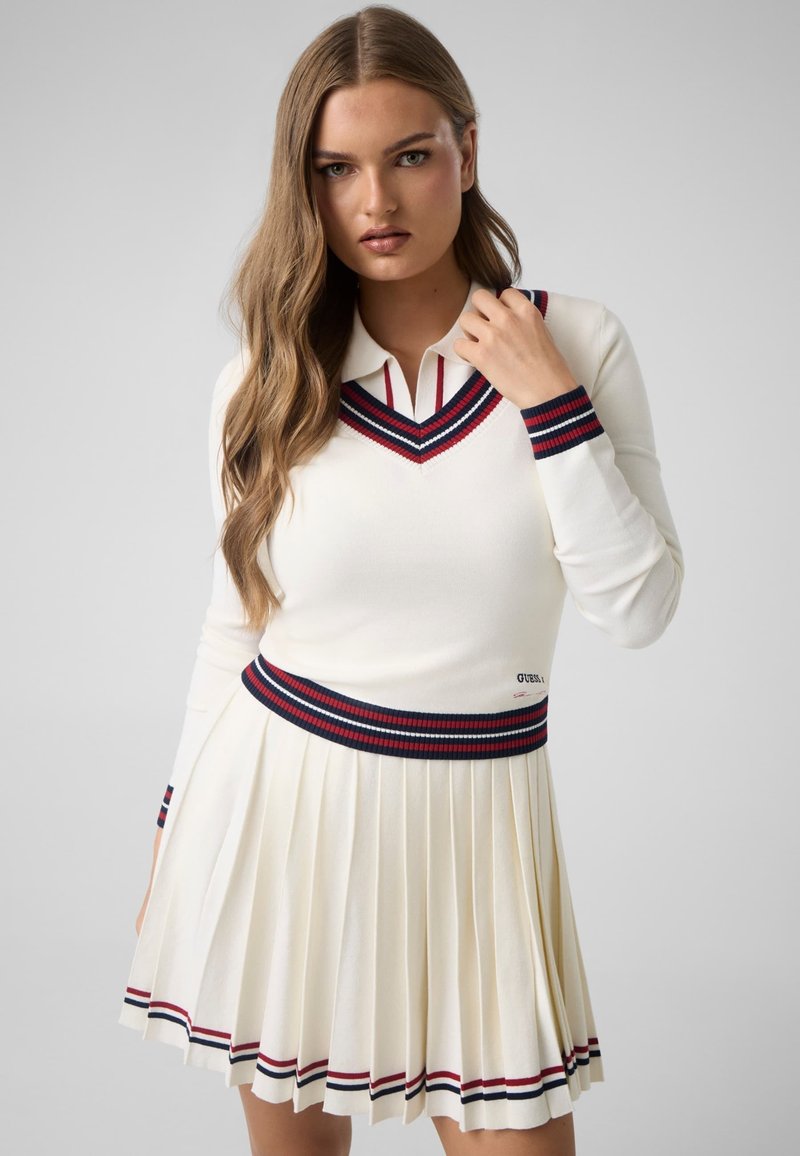 Femme aux longs cheveux portant une jupe plissée blanche et un pull assorti avec des bordures rayées rouges et bleu marine, posant devant un fond uni.