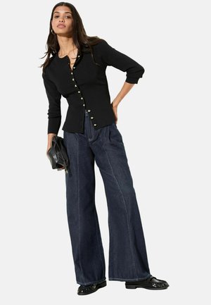 Schwarzer Knopfstrickcardigan mit Dreiviertelärmeln, kombiniert mit dunkelblauen Jeans mit weitem Bein. Das Model hält eine schwarze Lederclutch.