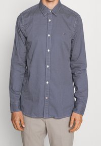 Camicia blu navy con pois bianchi, colletto all'ingiù e orlo arrotondato. Presenta bottoni frontali e un piccolo dettaglio con logo. Materiale in cotone.