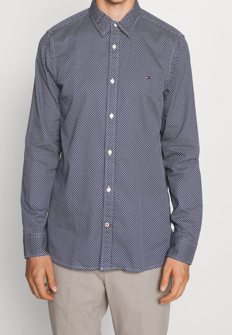 Camicia blu navy con pois bianchi, colletto all'ingiù e orlo arrotondato. Presenta bottoni frontali e un piccolo dettaglio con logo. Materiale in cotone.