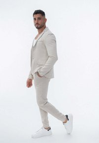 Costume beige clair avec une coupe ajustée, veste à un seul bouton, pantalon assorti et baskets blanches avec des accents noirs. Texture du tissu lisse.