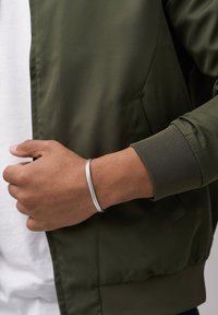 Hopeakukkavi bracelet kädessä, jossa on teksturoitu pinta, pukeutuneena vihreän pommirakenteen takin päälle, jossa on ribbikuviointi, valkoisen paidan päällä.