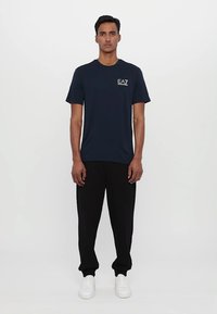 Námořnická modrá trička s jemným logem na hrudi, v kombinaci s černými kalhotami a bílými teniskami. Materiál se zdá být hladký, krátké rukávy, klasický střih.