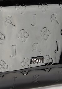Borsa in pelle verniciata nera con motivi floreali in rilievo e iniziali. Logo "JOOP!" in argento ben visibile sulla parte frontale. Texture liscia e lucida.