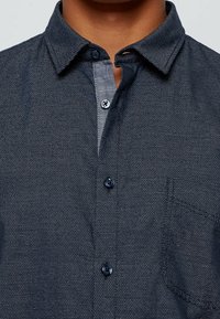 Mörkblå, texturerad skjorta med knäppning och klassisk krage, med kontrasterande ljusare tyg vid knäppningen och mörka knappar.
